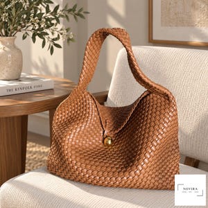 May include: A tan woven hobo bag featuring a single integrated shoulder strap and a central gold-toned spherical button closure. The exterior displays a consistent basket-weave pattern throughout the body and strap, with the brand name NOVIRA printed in the corner.