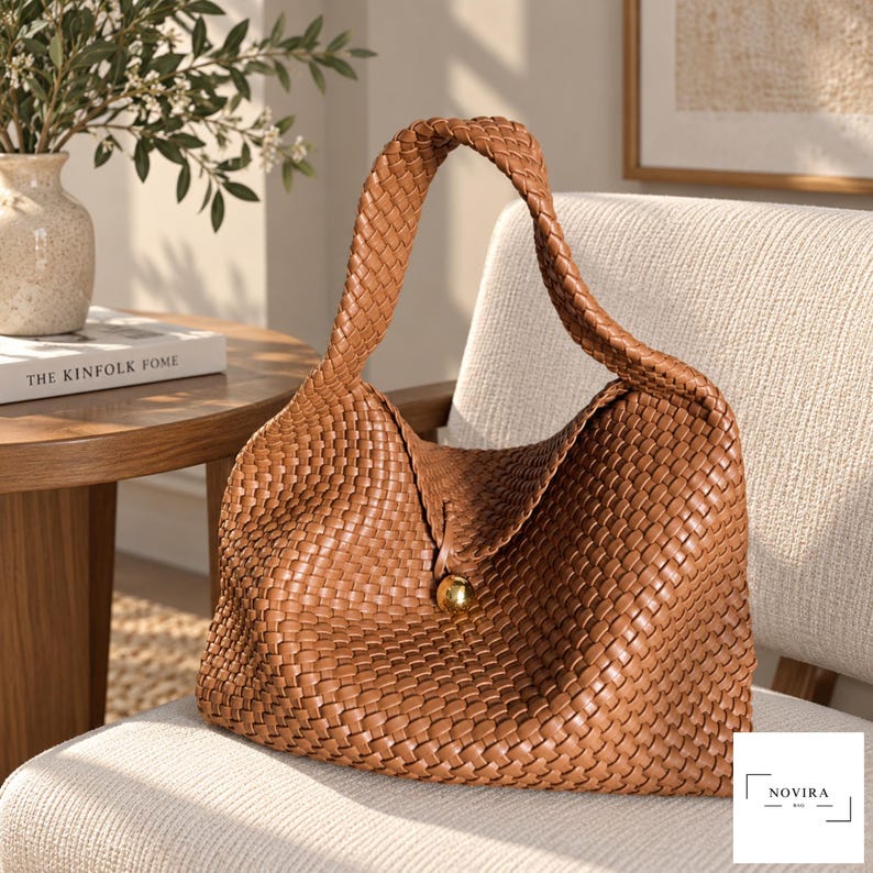 May include: A tan woven hobo bag featuring a single integrated shoulder strap and a central gold-toned spherical button closure. The exterior displays a consistent basket-weave pattern throughout the body and strap, with the brand name NOVIRA printed in the corner.