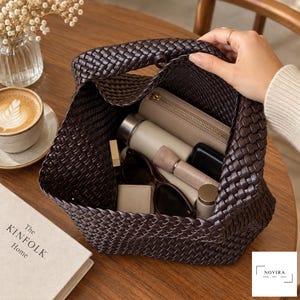 May include: A dark brown woven handbag with an open top, containing a beige zippered wallet, a cylindrical bottle, a small black case, a lipstick, and dark-rimmed sunglasses. The bag features a consistent braided pattern throughout its exterior.