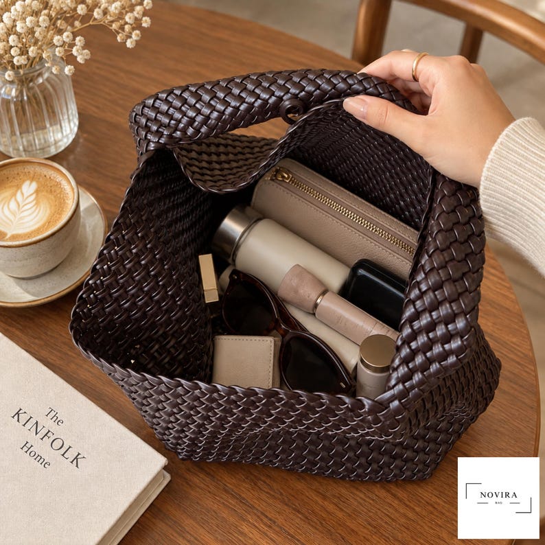 May include: A dark brown woven handbag with an open top, containing a beige zippered wallet, a cylindrical bottle, a small black case, a lipstick, and dark-rimmed sunglasses. The bag features a consistent braided pattern throughout its exterior.