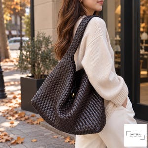 May include: A dark brown hobo bag featuring an intricate woven leather texture and a single shoulder strap. The bag includes a central gold-toned metal hardware accent and a small white logo tag in the corner with the text NOVIRA BAG.