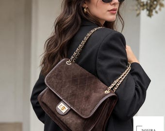 May include: A dark brown suede shoulder bag featuring a quilted diamond pattern, a gold-tone rectangular turn-lock closure, and a gold-tone chain strap interwoven with matching brown material. The bag includes a logo tag reading NOVIRA BAG.