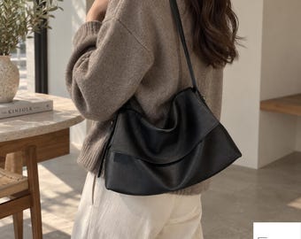 May include: A black shoulder bag with a smooth, matte finish and a structured, slightly slouchy silhouette. The bag features a single long strap and a fold-over flap design. The bottom corner displays the text NOVIRA BAG.