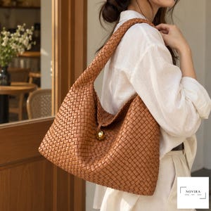 May include: A tan hobo bag featuring an intricate woven leather pattern, a single wide shoulder strap, and a central gold-toned spherical clasp closure. The bag displays the text NOVIRA BAG in a rectangular frame in the bottom right corner.