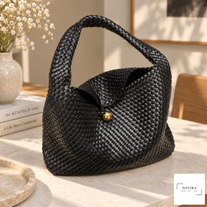 May include: A black woven handbag featuring a structured hobo silhouette with a single integrated handle. The bag is secured with a central gold-toned spherical button closure. The exterior surface displays a consistent, intricate basket-weave pattern.