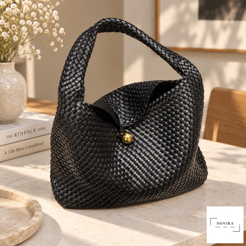 May include: A black woven handbag featuring a structured hobo silhouette with a single integrated handle. The bag is secured with a central gold-toned spherical button closure. The exterior surface displays a consistent, intricate basket-weave pattern.