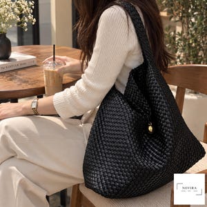 May include: A black woven hobo bag featuring a textured, interlaced pattern and a single shoulder strap. The bag includes a small, round gold-toned metal charm hanging from the side. The design is structured with a soft, slouchy silhouette.