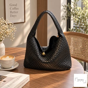 May include: A black woven hobo bag featuring a single integrated shoulder strap and a central gold-toned spherical charm. The bag is constructed from a dense, uniform basket-weave pattern, creating a textured surface throughout the entire exterior.