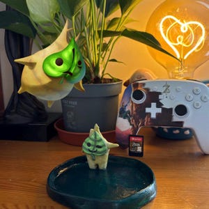 May include: Two small, light yellow, leaf-shaped figurines with green leaf masks, one floating and one standing on a dark green circular tray. Next to them is a white game controller with a character graphic and a small rectangular game cartridge.