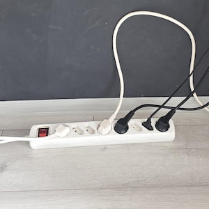 Op de afbeelding: Een witte stekkerdoos met een rode aan-uitknop, voorzien van meerdere aangesloten zwarte en witte stroomkabels. De stekkerdoos is verbonden met een stopcontact via een wit snoer, waarbij diverse andere zwarte kabels omhoog lopen vanaf de unit.