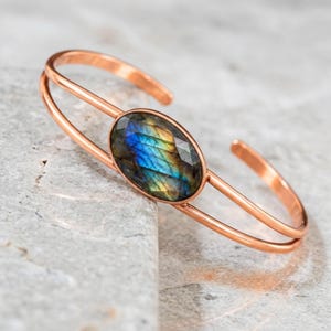 May include: A copper cuff bracelet featuring a central oval-shaped labradorite gemstone. The stone displays iridescent flashes of blue and gold. The bracelet is constructed from two parallel copper bands that meet at the stone setting.