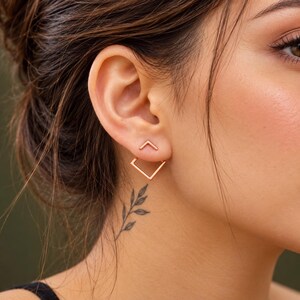 May include: A rose gold ear jacket earring featuring a small chevron-shaped stud at the front of the earlobe and a larger, open square-shaped frame that hangs below the earlobe, creating a geometric layered effect on the ear.