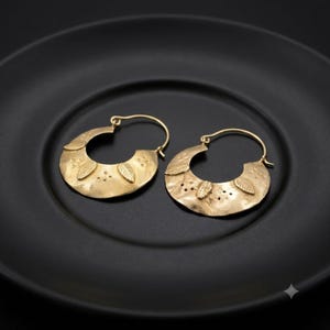 May include: A pair of gold-toned hoop earrings with a wide, crescent-shaped design. The surface of each earring features a textured, hammered finish with embossed leaf motifs and small dot patterns along the curved metal band.