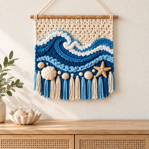 May include: A macrame wall hanging featuring a cream-colored top section and layered waves in shades of blue. The piece is adorned with crocheted white foam, various seashells, and a starfish, finished with alternating cream and blue yarn tassels.