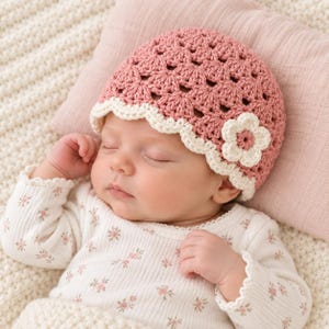 May include: A dusty rose crochet baby hat featuring a scalloped cream-colored border and a single cream-colored flower appliqué on the side. The hat is crafted with a textured shell stitch pattern and is worn by a sleeping infant in a floral top.