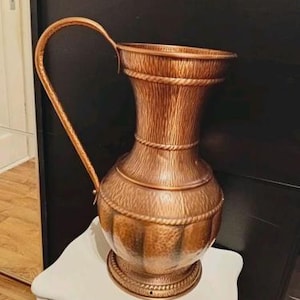 Op de afbeelding: Een hoge koperen kan met een getextureerde, gehamerde afwerking met verticale groeven op het bolvormige lichaam en de hals. De kan is voorzien van een gebogen handvat, een uitlopende rand en decoratieve touwachtige details rond de hals en voet.