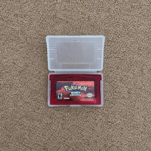 Puede incluir: Un cartucho de videojuego rojo translúcido para Game Boy Advance titulado Pokémon Ruby Version, alojado dentro de una funda protectora de plástico transparente. La etiqueta presenta el logotipo de Pokémon, el texto Ruby Version y un símbolo E.