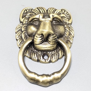 Può includere: Un battente per porta in metallo color ottone raffigurante una testa di leone scolpita con criniera e tratti del viso dettagliati. Un anello circolare è sospeso alla bocca del leone e un piccolo foro di montaggio è visibile sotto il mento.