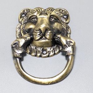 May include: A brass-coloured metal door knocker featuring a central lion head design with a circular ring held by two smaller animal heads on either side. The piece includes visible screw holes at the top and bottom for mounting to a flat surface.