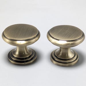May include: Two round cabinet knobs with a brushed antique brass finish. Each knob features a flat circular top, a tapered neck, and a stepped circular base. The metal surface displays a subtle horizontal grain texture throughout the design.