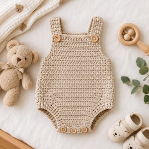 May include: A beige crochet baby romper featuring a sleeveless design with two wooden buttons on the front straps and three wooden buttons at the bottom leg opening. The garment is crafted with a textured stitch pattern in a neutral tan color.