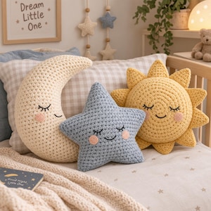 Puede incluir: Tres cojines de ganchillo con forma de media luna, estrella de cinco puntas y sol. La luna es color crema, la estrella es azul claro y el sol es amarillo. Cada cojín presenta ojos cerrados bordados, una sonrisa y mejillas rosadas.