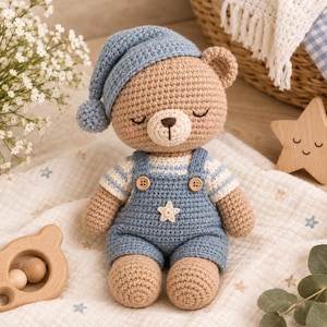 Könnte beinhalten: Ein gehäkelter Teddybär aus beigem Garn, der eine blaue Schlafmütze mit Bommel, ein weiß-blau gestreiftes Shirt und eine blaue Latzhose mit zwei Holzknöpfen sowie einer kleinen weißen Sternapplikation auf der Brust trägt, mit geschlossenen Augen.