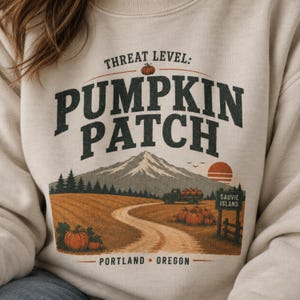 May include: A beige crewneck sweatshirt featuring a graphic print with the text THREAT LEVEL: PUMPKIN PATCH, PORTLAND OREGON, and SAUVIE ISLAND. The illustration depicts a pumpkin field, a mountain, a truck, and a sunset in orange and green.