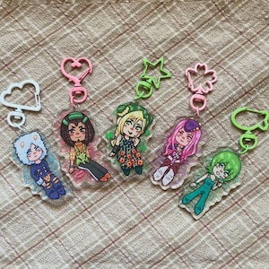 May include: Five acrylic keychains featuring stylised anime-inspired characters. Each charm has a unique coloured plastic clasp: white cloud, pink heart, green star, pink flower, and green cat shape. The characters have distinct hair and clothing.