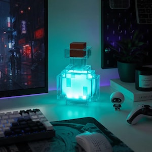 May include: A square, pixelated potion bottle lamp with a clear, blocky structure and a brown stopper. The interior glows with a bright, vibrant cyan light, casting a soft illumination on the surrounding desk area in a gaming setup.
