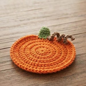 Könnte beinhalten: Ein runder, orangefarbener Häkeluntersetzer in Kürbisform. Der Untersetzer weist ein spiralförmiges Stichmuster, einen kleinen grünen gehäkelten Stiel in der oberen Mitte und eine braune, lockige gehäkelte Ranke an der rechten Seite des Stiels auf.