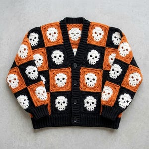 May include: A long-sleeve crochet cardigan featuring a checkerboard pattern of black and orange squares, each containing a white skull motif. The garment has a V-neckline, a black ribbed trim along the front opening, and four black buttons.