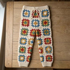 May include: Crochet pants constructed from individual granny squares in cream, blue, yellow, red, and green. The pants feature a ribbed elastic waistband with a matching cream-colored drawstring tie and ribbed cuffs at the bottom of each leg.