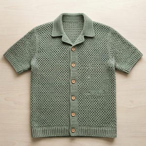 Könnte beinhalten: Ein kurzärmeliges Hemd in Salbeigrün mit einem gehäkelten Strickmuster. Das Kleidungsstück zum Knöpfen verfügt über einen klassischen Kragen, eine Knopfleiste mit sechs hellbraunen Holzknöpfen sowie gerippte Abschlüsse an Ärmeln und Saum.