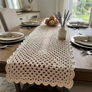 Könnte beinhalten: Ein rechteckiger, cremefarbener Häkel-Tischläufer mit einem sich wiederholenden Lochmuster aus Granny Squares und einem gewellten Rand. Der Läufer liegt flach auf einem Holztisch, zentriert unter einer Keramikvase mit getrocknetem Lavendel.
