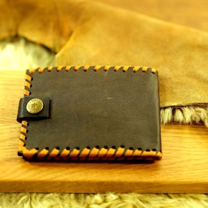 Op de afbeelding: Een donkerbruine lederen bifold portemonnee met een opvallende oranje festonsteek langs de randen. De portemonnee heeft een kleine leren sluiting met een ronde, messingkleurige metalen drukknoop voorzien van een ster en tekst.