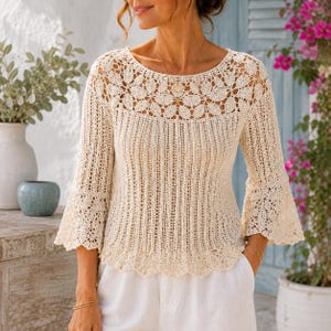 May include: A cream-colored crochet top featuring a round neckline with an intricate floral pattern, long bell sleeves, and a vertical ribbed knit texture throughout the bodice. The garment has a scalloped hemline and is crafted from lightweight yarn.