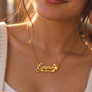 Könnte beinhalten: Eine goldfarbene Halskette mit einem Namensanhänger, der den Namen Emma in Schreibschrift zeigt. Der Anhänger ist auf der linken Seite mit einem kleinen Herz und auf der rechten Seite mit drei fünfzackigen Sternen in verschiedenen Größen verziert.