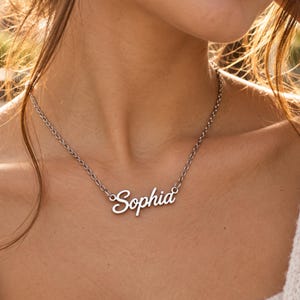 Könnte beinhalten: Eine silberfarbene Namenskette aus Metall mit dem Namen Sophia in einer kursiven Schriftart. Der Anhänger ist an einer zarten Kette mit ovalen Gliedern befestigt, die um den Hals liegt. Das Design ist schlicht und hat eine polierte Oberfläche.