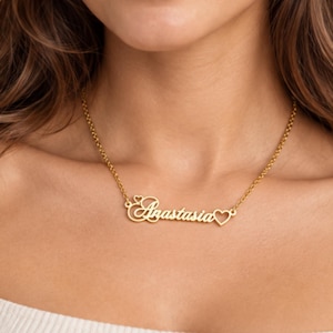 Könnte beinhalten: Eine goldfarbene Halskette mit einem Namensanhänger in Schreibschrift, der den Namen Anastasia zeigt. Der Name wird von zwei kleinen herzförmigen Aussparungen flankiert, eine über dem ersten Buchstaben und eine am Ende des Namens, befestigt an einer Kette.