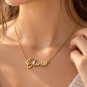 Könnte beinhalten: Eine goldfarbene Metallhalskette mit einem Namensanhänger, auf dem der Name Olivia in einer kursiven Schriftart steht. Der Anhänger ist an einer zarten, dünnen Gliederkette befestigt, die um den Hals liegt und eine polierte Oberfläche aufweist.