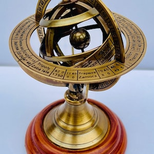 May include: A brass armillary sphere featuring a central globe and rotating rings marked with degrees, city names like Rouen, Tripoli, and Zamora, and the zodiac sign Sagittarius, all mounted on a circular, polished reddish-brown wooden base.