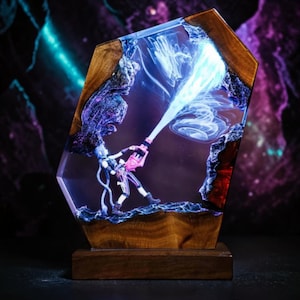 Op de afbeelding: Een decoratieve lamp met een miniatuurfiguur met blauw haar die een roze wapen vasthoudt dat een helderblauwe lichtstraal uitstraalt. Het figuur is ingesloten in een helder harsblok met een donkerhouten frame en een wervelend lichteffect binnenin.
