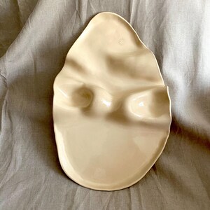 Pode incluir: Uma travessa de cerâmica de formato orgânico irregular com acabamento em creme brilhante. A superfície apresenta duas reentrâncias ou cavidades lisas e distintas moldadas no centro, com uma pequena marca circular do fabricante estampada perto da borda superior.