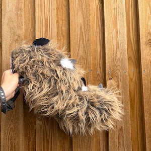 Può includere: Un accessorio in pelliccia sintetica marrone e marrone chiaro, caratterizzato da macchie bianche e una base in tessuto nero. L'articolo include un cinturino regolabile grigio scuro con componenti in metallo, progettato per essere indossato o fissato.