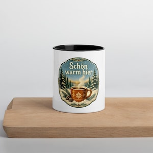 May include: A white ceramic mug with a black rim featuring a circular graphic of a steaming brown mug with a snowflake pattern, surrounded by pine trees and snow, with the text "Schön warm hier" printed in a decorative font above the mug.