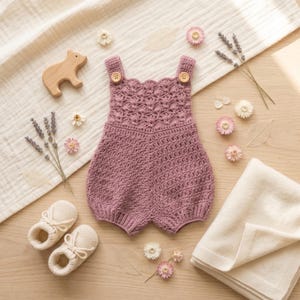 May include: A mauve crochet romper featuring a scalloped bodice, two wooden buttons at the shoulder straps, and a textured stitch pattern. The garment is displayed alongside a pair of cream-colored knit booties, a wooden bear figure, and dried flowers.
