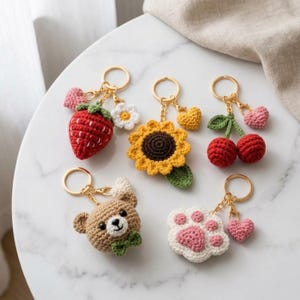 May include: Five crochet keychains featuring a red strawberry with a white flower, a yellow sunflower, two red cherries with a green leaf, a brown bear head with a green bow tie, and a white paw print with pink pads, each with a small heart charm.