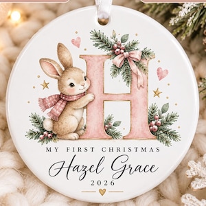 May include: A round white ceramic Christmas ornament featuring a brown bunny wearing a pink scarf next to a large pink letter H. The design includes pine branches, berries, and stars. Text reads: My First Christmas, Hazel Grace, and 2026.