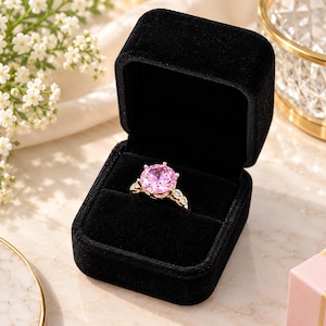 May include: A gold ring featuring a large, round, faceted pink center stone held by six prongs, with smaller clear stones set along the band, presented inside an open, square, black velvet jewelry box with a soft interior lining.